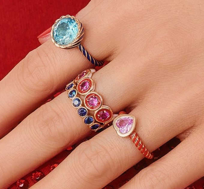 Close-up of a hand wearing three colorful gemstone rings on a red background