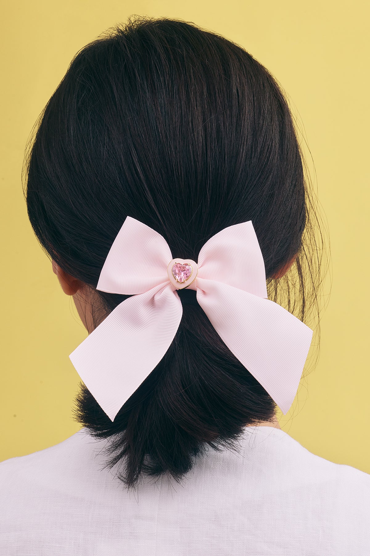 Pink Hair bow with pink crystal heart clipped on the back of models dark hair against yellow background