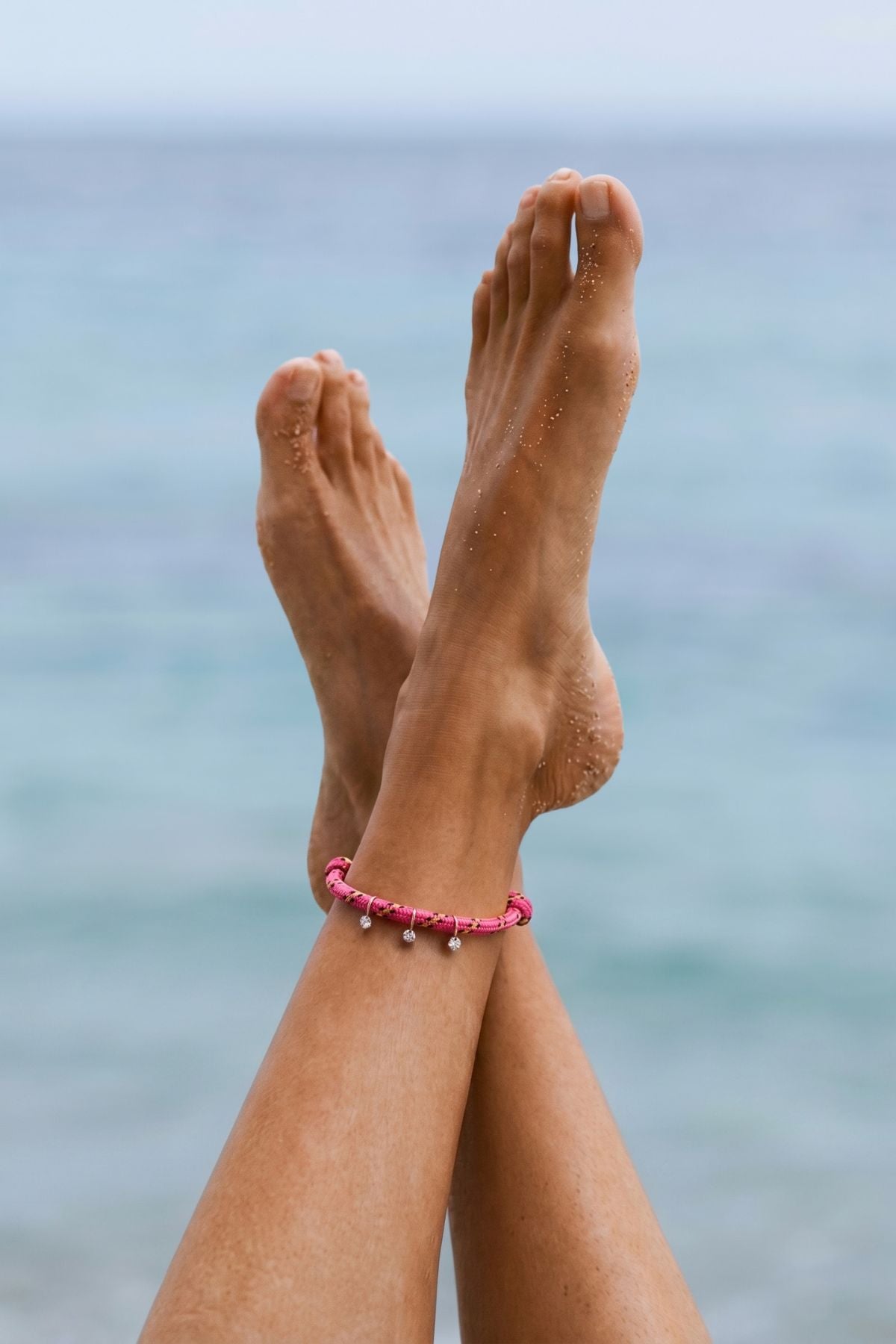 Model's feet with a pink anklet on her ankle against a blurred ocean background